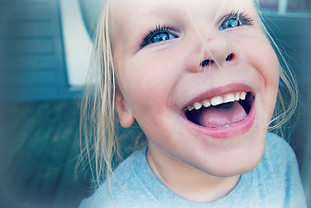 a young child with light blonde hair, bright blue eyes, and an open-mouthed smile, showing their teeth.