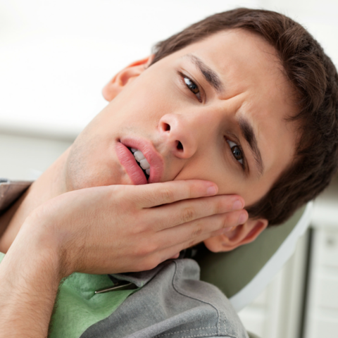 A young man in a dentist's chair, holding his cheek with a pained expression, indicating he is experiencing dental discomfort or toothache. The image captures the moment of distress, emphasizing the need for dental care.