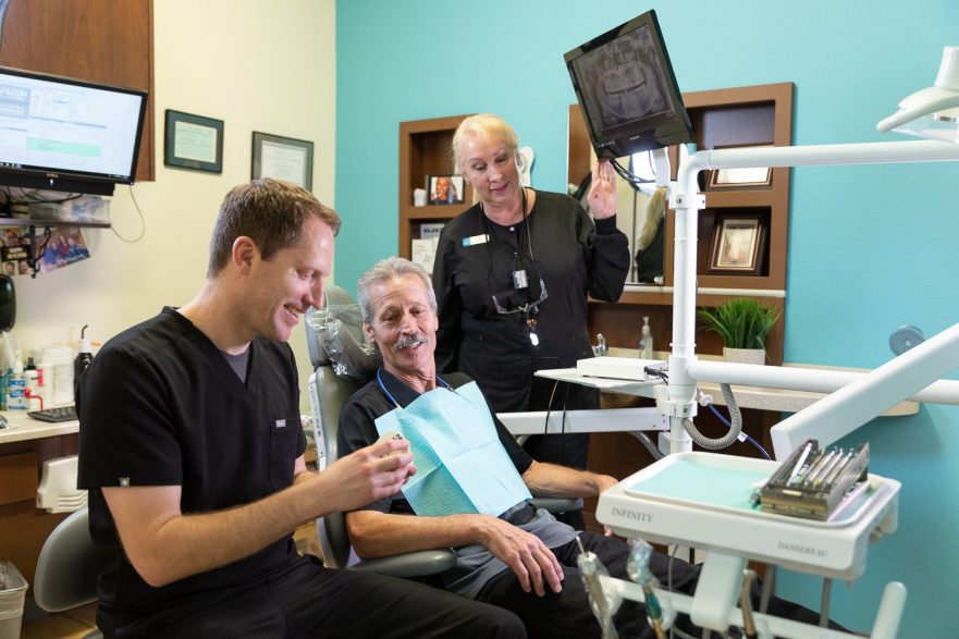 Dr. Mink, seated and smiling, holds a dental tool while explaining something to an older male patient who is seated in the dental chair, also smiling. A dental assistant stands nearby, attentively watching and holding a screen that displays an image of the patient’s dental X-ray. The room is equipped with various dental instruments, and the atmosphere appears to be friendly and professional.