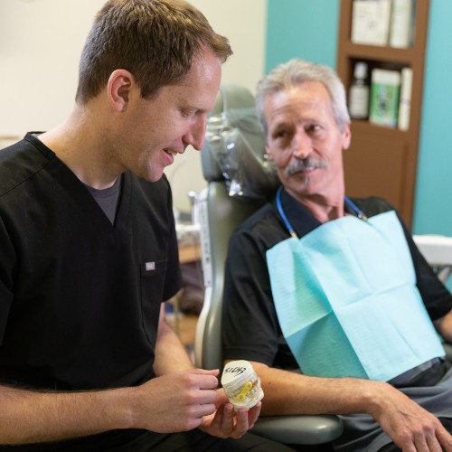 Dr. Mink, dressed in black scrubs, is seated next to an older male patient who is wearing a dental bib. Dr. Mink is holding a dental mold and explaining something to the patient, who is attentively listening. 