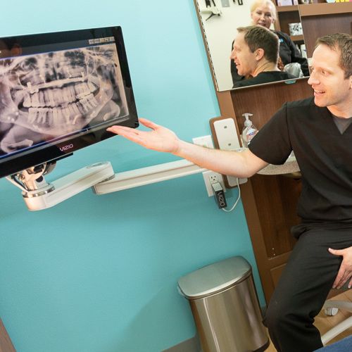  Dr. Mink, in black scrubs is pointing to an X-ray image displayed on a monitor. The X-ray shows a detailed view of a patient's teeth and jaw. The dentist is explaining something to a patient.