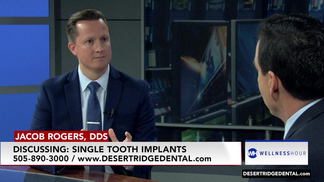 Dr. Jacob Rogers, DDS, during a television interview on the "Wellness Hour." Dr. Rogers is wearing a suit and tie, discussing single tooth implants. He is facing the host, who is partially visible from behind. The background includes various screens and a modern studio setting. The text at the bottom of the image reads, "Jacob Rogers, DDS, Discussing: Single Tooth Implants, 505-890-3000 / www.DESERTRIDGEDENTAL.com." The "Wellness Hour" logo is also displayed.