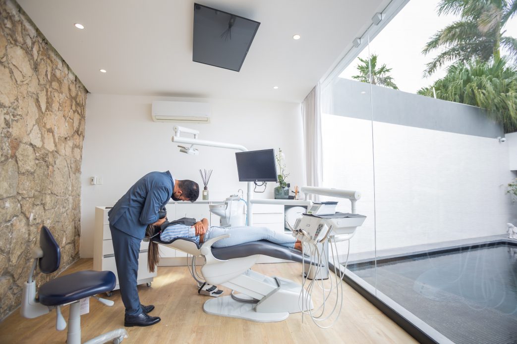a modern dental office with large windows and a view of an outdoor space with palm trees. A dentist is leaning over a patient who is lying on a dental chair. The room is bright and spacious, with a stone wall on one side and sleek dental equipment, including a monitor and overhead light.